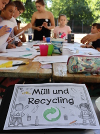 Kinder arbeiten an einem Tisch mit Papiermaterialien, davor liegt ein Infoblatt zum Thema Müll und Recycling